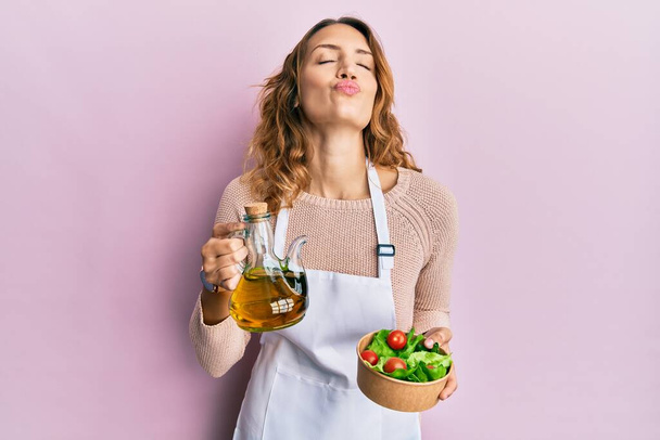 Fiatal kaukázusi nő kötényt visel, olívaolajos kannával és salátával a kamerába néz, és egy csókot fúj, hogy kedves és szexi legyen. szerelem kifejezés.  - Fotó, kép