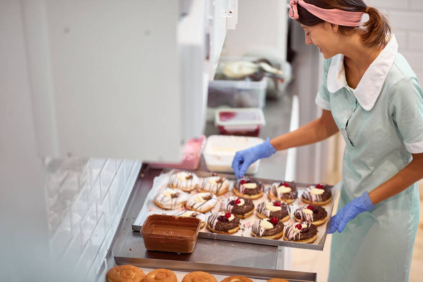 A young small business female owner is decorating handmade delicious donuts with a passion in a relaxed atmosphere in a workshop. Pastry, dessert, sweet, making - Photo, Image