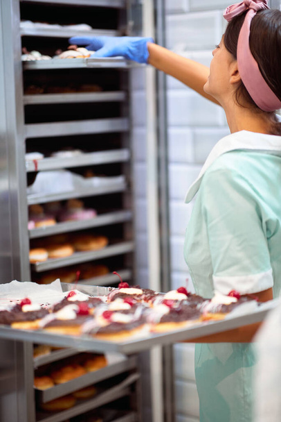 A young small business female owner is taking handmade delicious donuts out from an owen in a working atmosphere in a candy workshop. Pastry, dessert, sweet, making - Photo, Image