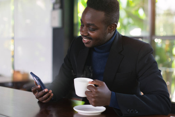 Empresario africano beber un café caliente en la cafetería con noticias en busca de teléfonos inteligentes - Foto, Imagen