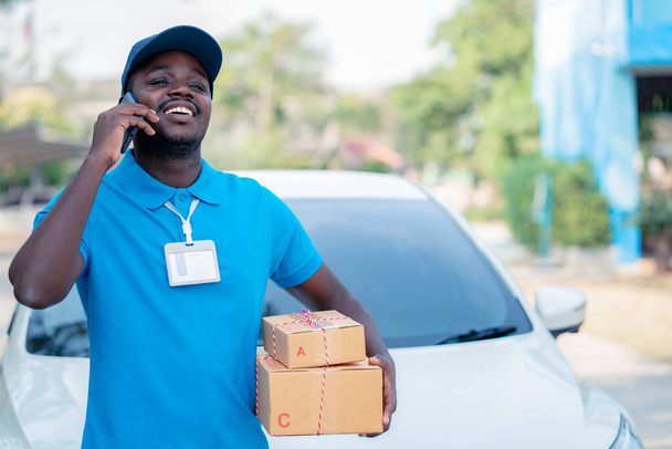 Hombre africano mensajero mensajero de entrega postal que sostiene la entrega de cajas de paquetes delante del coche con el teléfono inteligente que habla - Foto, imagen