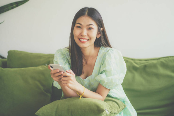 Kaunis nuori hymyilevä nainen käyttää matkapuhelintaan istuu pohjoismainen vihreä sohva - Valokuva, kuva