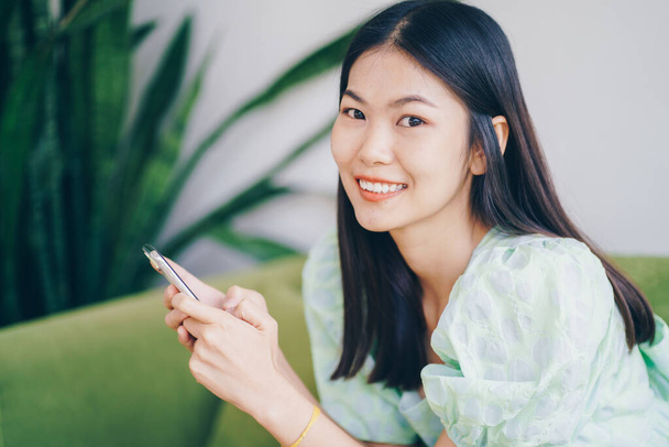 Досить молода усміхнена жінка використовує свій мобільний телефон, сидячи на скандинавському зеленому дивані
 - Фото, зображення