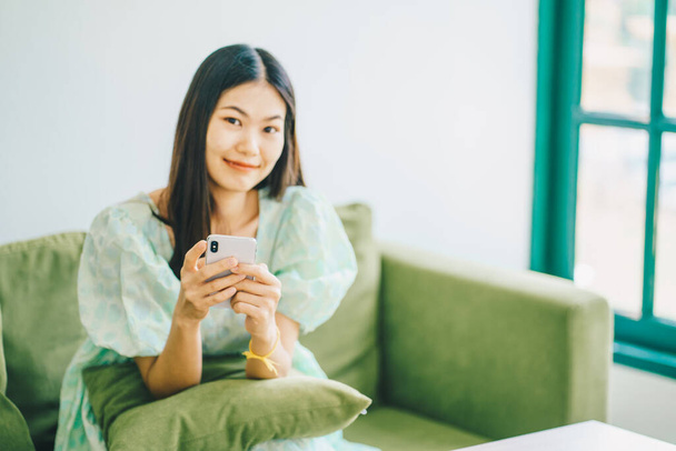 Pretty young smiling woman using her mobile phone sitting on nordic green sofa - Photo, Image