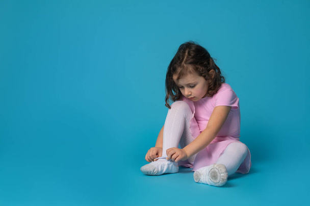 Cute ballerina focused on tying shoelaces on ballet shoes sitting on blue background with copy space - 写真・画像