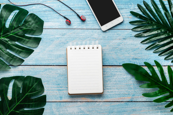 Boş not defteri, telefon, kulaklık mavi ahşap kopya alanı üzerinde tropikal yeşil yapraklar. Üst görünüm - Fotoğraf, Görsel
