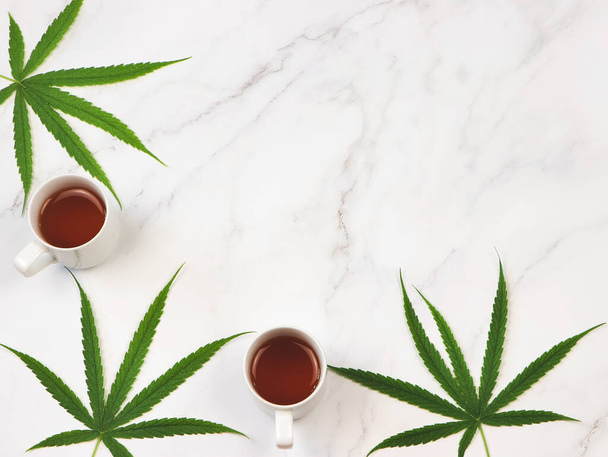 Vista superior o plano de dos tazas de té de cannabis y hojas de marihuana sobre fondo de mármol blanco. - Foto, imagen