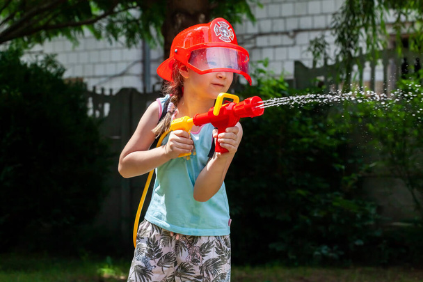 消防士、幼い子供、庭で水鉄砲で遊ぶ消防士のヘルメットを身に着けている小さな学齢の女の子。夢、将来の職業、仕事、遊び、楽しいアウトドアライフスタイルのコンセプト - 写真・画像