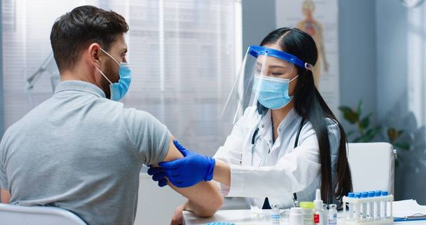 Close up portrait of pretty Asian female doctor in medical mask and face shield working in clinic cabinet during vaccination to handsome Caucasian male patient. Covid pandemic, vaccine cure concept - Foto, immagini