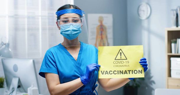 Lähikuva muotokuva Aasian nuori terveydenhuollon työntekijä sairaanhoitaja suojaava kasvot kilpi ja naamio pitämällä käsissä käsineet kortti sanomalla Covid-19 rokotus. ilmoitus sepelvaltimotautirokotteesta - Valokuva, kuva