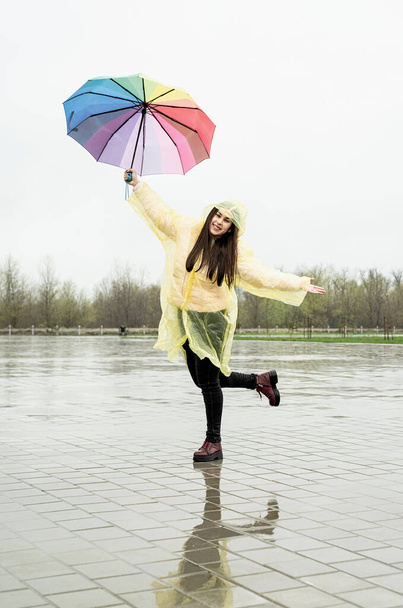 Красивая брюнетка в жёлтом плаще с радужным зонтиком танцует под дождем - Фото, изображение