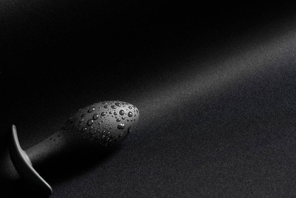A black butt plug with drops of moisture lies on a dark surface. Friction diagonal and direction. Sex toys and sex pleasure concept. Close-up - Photo, image