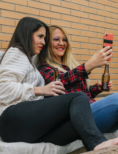 Due giovani amici sorridenti che bevono birra e scattano selfie con un cellulare su una terrazza - Foto, immagini