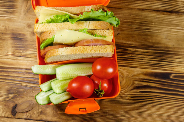 Обеденная коробка со сэндвичами и свежими овощами на деревянном столе. Вид сверху - Фото, изображение