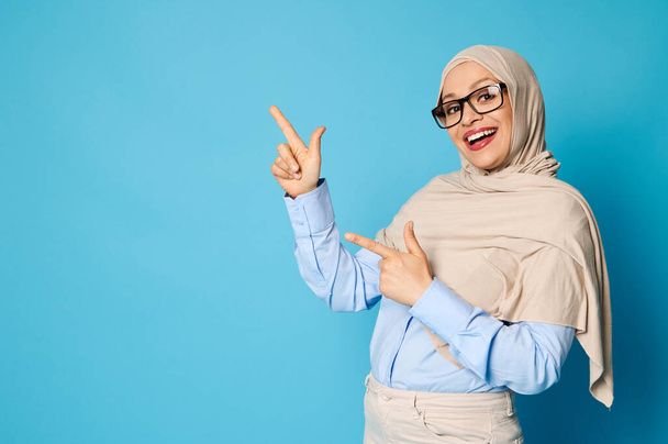 Okouzlující arabská žena v hidžábu s krásným úsměvem ukazuje na její ukazováček na modrém pozadí s kopírovacím prostorem - Fotografie, Obrázek