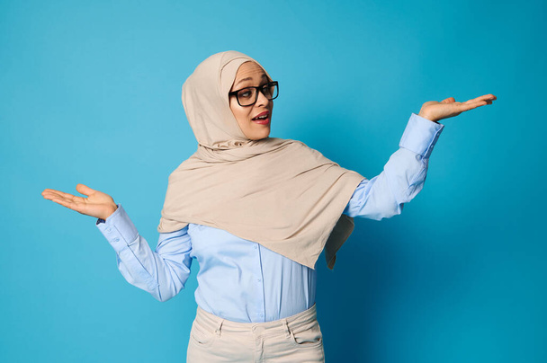 Araberin im Hijab posiert mit ausgestreckten Armen und nach oben gestreckten Handflächen, präsentiert auf blauem Hintergrund mit Platz für Text - Foto, Bild