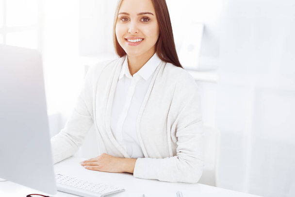 Young cheerful woman sitting at the desk with computer and looking at camera in sunny office. Headshot - Фото, зображення