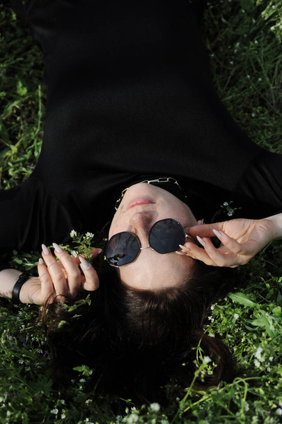 Beautiful young woman in sunglasses enjoying the spring. Woman lying in the grass outdoors. - Foto, afbeelding