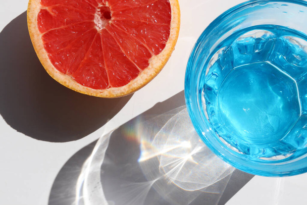 Летний натюрморт с голубым стаканом холодной воды и половиной грейпфрута на белом фоне стола. Вид сверху, место для копирования. Жесткий свет, тень - Фото, изображение