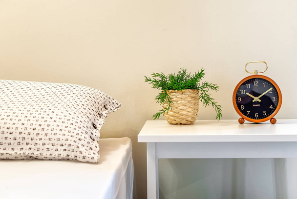 Table de chevet avec plante verte dans un pot de paille et horloge dessus. Une partie du lit est visible. Détails intérieurs. couleurs claires.  - Photo, image