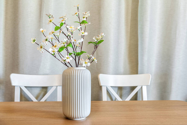 Jarrón blanco con ramas con flores en él, de pie sobre la mesa ligera de madera. Enfoque selectivo. Parte de las sillas en el fondo borroso - Foto, Imagen