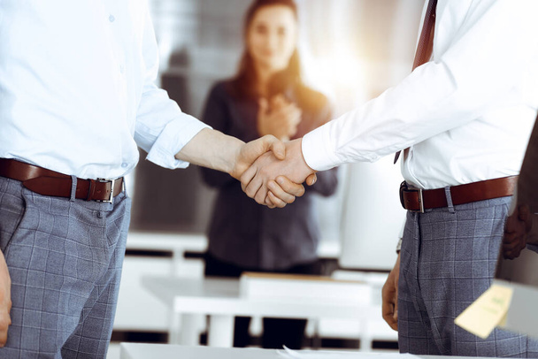 Two businessmen shaking hands in sunny office, close-up. Happy and excited business woman stands with raising hands at the background. Business people concept - Photo, image