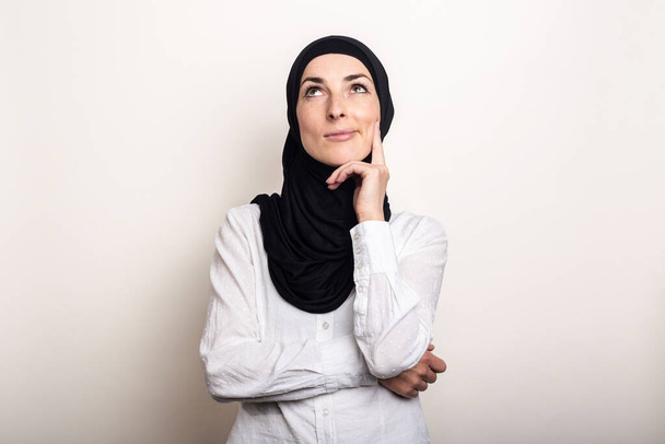 Egy fiatal nő fehér ingben és hidzsáb tartja a kezét az állához, és felnéz egy töprengő arccal, világos háttérrel. Zászlós. - Fotó, kép