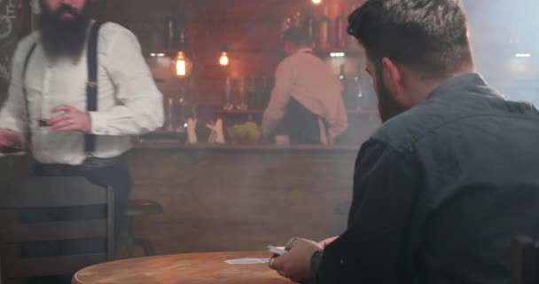 Γενειοφόρος άνθρωπος αρπάξει ποτά από το μπαρ και ξεκινά ένα παιχνίδι καρτών με το φίλο του - Πλάνα, βίντεο