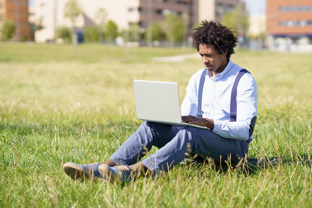 Homme noir aux cheveux afro utilisant son ordinateur portable assis sur une planche à roulettes sur l'herbe d'un parc urbain. - Photo, image