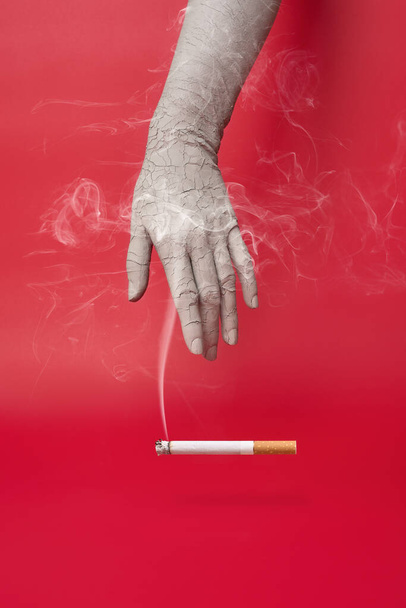 Mano seca y agrietada y un cigarrillo humeante sobre fondo rojo. - Foto, imagen