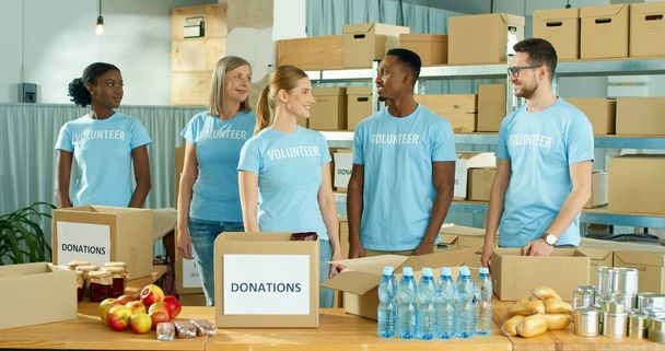 Multi-ethnic cheerful diverse men and women social workers standing in charity organization warehouse with donations, looking at camera and smiling. Charity volunteers work. Donating and volunteering - 写真・画像