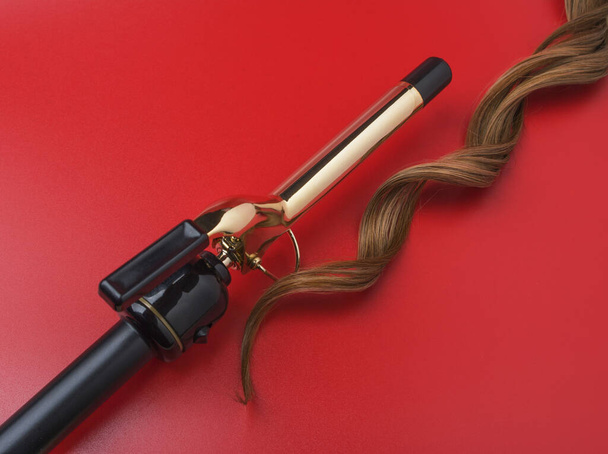Professional Hairdressing Curling Iron With A lock Of Waved Hair on A Red Background, Spotted With Ray Of Lights - Photo, Image