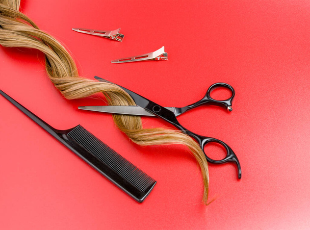 Otevřené kadeřnické nůžky s pramenem kudrnaté vlasy na červeném pozadí, profesionální nůžky, klipy a ocas hřeben - Fotografie, Obrázek