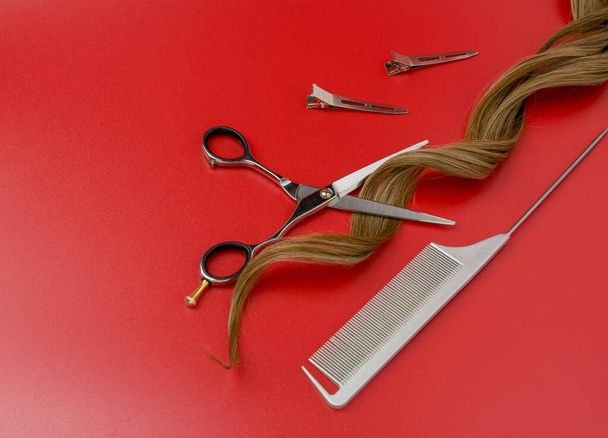 Abrir tesoura de cabeleireiro com uma mecha de cabelo encaracolado artificial em fundo vermelho. Tesouras profissionais, pente de prata e clipes de metal - Foto, Imagem