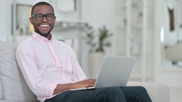 Uomo africano con computer portatile sorridente alla fotocamera a casa  - Foto, immagini