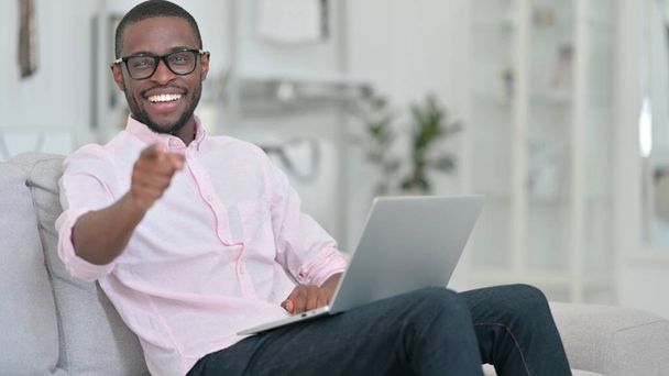Emocionado hombre africano con el ordenador portátil señalando a la cámara en casa
  - Foto, imagen