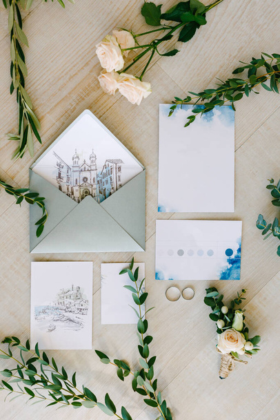 Svatební prsteny leží na dlaždicové podlaze, obklopené akvarelovými malbami, zelenými větvemi a květinami. Obrazy zobrazují staré domy - Fotografie, Obrázek