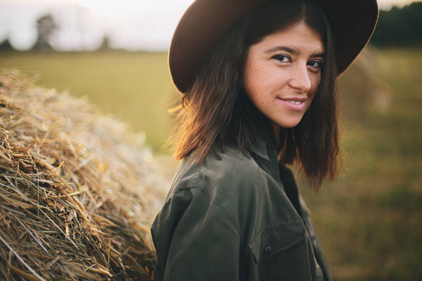 Beautiful stylish woman in hat standing at hay bale in summer evening field. Atmospheric portrait of young fashionable female relaxing at haystacks, summer vacation in countryside - Photo, image