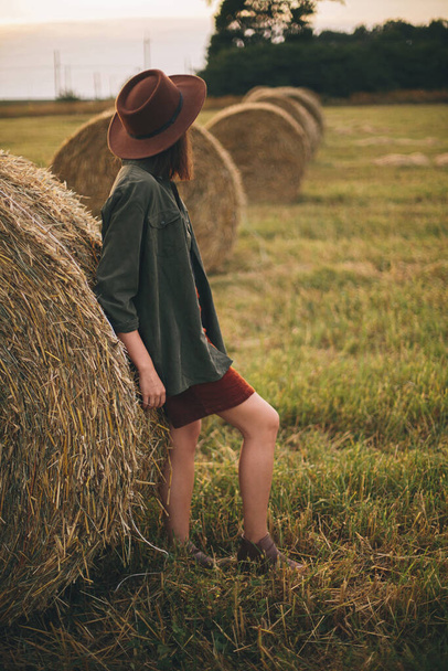 Stylish woman in hat standing at hay bale in summer evening in field. Atmospheric tranquil moment. Young fashionable female relaxing at haystack, summer vacation in countryside - Photo, Image