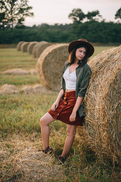 Tyylikäs hattuinen nainen seisoo heinäpaalilla kesäiltana pellolla. Ilmakehän rauhallinen hetki. Nuori muodikas nainen rentouttava heinäsuovassa, kesäloma maaseudulla. Maaseudun hidas elämä - Valokuva, kuva