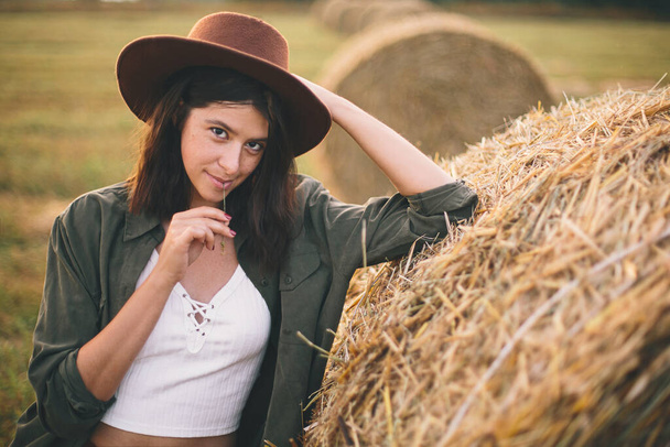 Beautiful stylish woman with straw in mouth and in hat posing at haystacks in summer evening field. Sexy young female relaxing at hay bale, summer vacation in countryside. - Φωτογραφία, εικόνα
