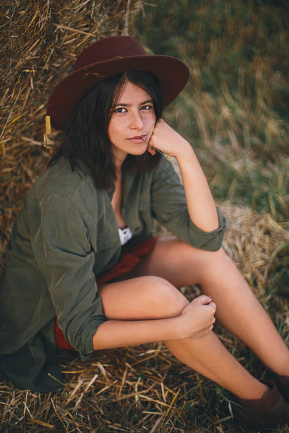 口の中にハーブと夏の夜のフィールドで干し草の山に座って帽子を持つ美しいスタイリッシュな女性。セクシーな若い女性の肖像画は、田舎で夏休み、干し草のベールでリラックス. - 写真・画像