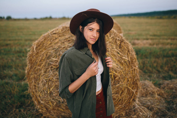 帽子の美しいスタイリッシュな女性は夏の夜のフィールドで干し草の俵に立っています。若いファッショナブルな女性の肖像画は、田舎の夏休み、干し草でリラックス。静けさ - 写真・画像