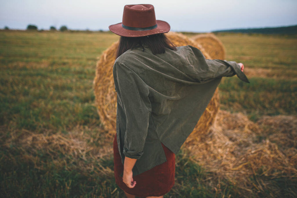 Стильная женщина в шляпе, танцующая у сенокосов на летнем вечернем поле. Беззаботный счастливый момент, отдых за городом. Счастливая молодая женщина расслабляется и веселится у стогов сена - Фото, изображение