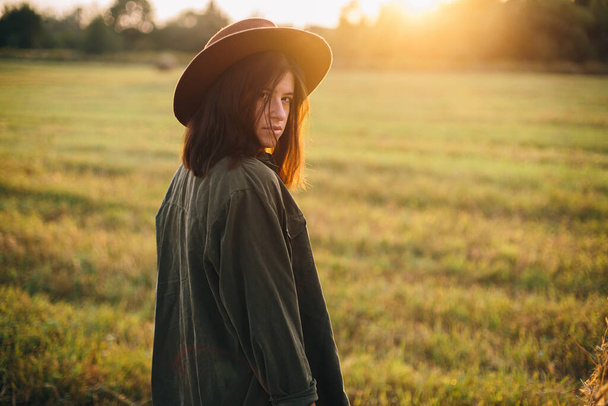 Стильная женщина в шляпе, позирующая у стогов сена при солнечном свете в летнем поле. Атмосферный спокойный момент в сельской местности. Молодая женщина отдыхает на сене в вечернее теплое солнце - Фото, изображение