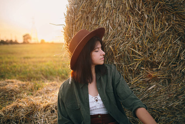 帽子の美しいスタイリッシュな女性は、日当たりの良い夏のフィールドで夜を楽しむ干し草に座っています。田舎の静かな瞬間。若いです女性リラックスで干し草のベールで暖かい日没 - 写真・画像