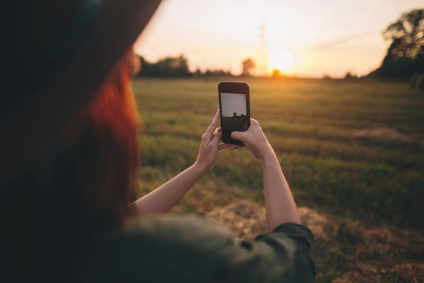 Stylowa kobieta robi zdjęcie zachodu słońca przez telefon na letnim polu. Młoda kobieta trzyma smartfona i przechwytuje wieczorne ciepłe słońce na wsi. Atmosferyczna piękna chwila - Zdjęcie, obraz