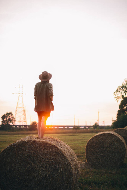 Beautiful carefree woman in hat standing on haystack enjoying evening sunset in summer field. Young happy female relaxing on hay bale in countryside. Atmospheric tranquil moment - Photo, Image
