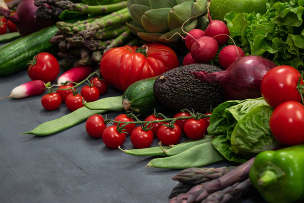 Különböző organikus zöldségek - spárga, paradicsom cseresznye, avokádó, articsóka, bors, lime, citrom, só szürke alapon. Nyers vegetáriánus fogalom. - Fotó, kép