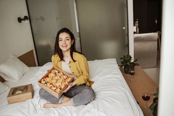 Милая женщина, сидящая на кровати с коробками с едой, полными круассанов, имеет романтический ужин дома - Фото, изображение